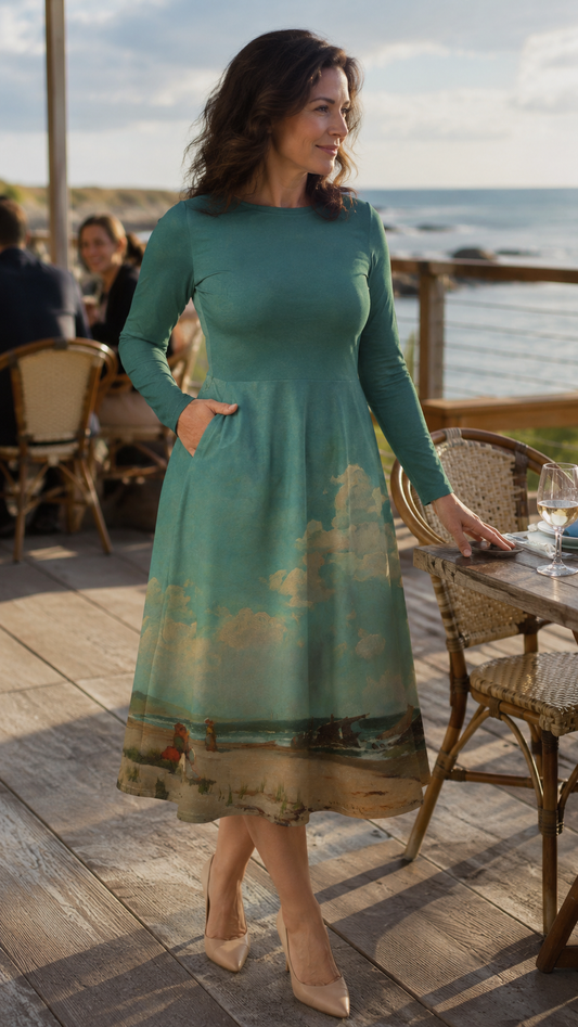 A woman wearing a teal long-sleeve midi dress with a beach landscape print at the hem, standing on a wooden deck at an outdoor cafe near the ocean.