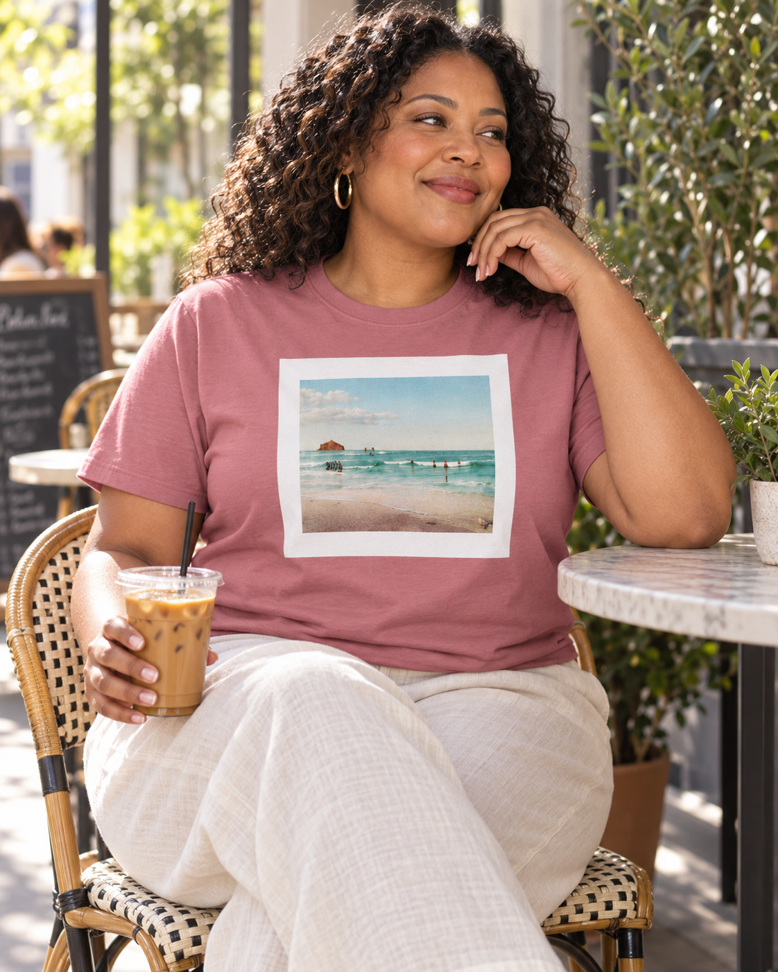A woman sitting at an outdoor cafe table wearing a dusty rose t-shirt with a square beach graphic print on the front, paired with light beige pants.