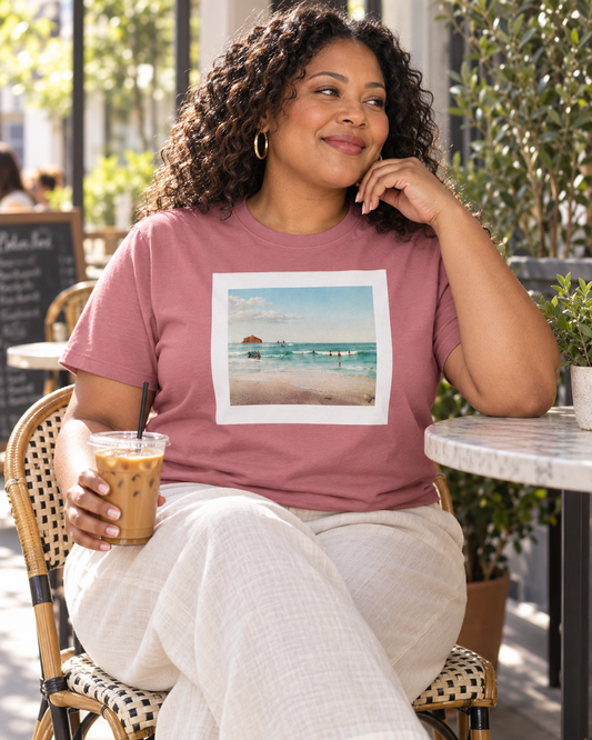 A woman sitting at an outdoor cafe table wearing a dusty rose t-shirt with a square beach graphic print on the front, paired with light beige pants.