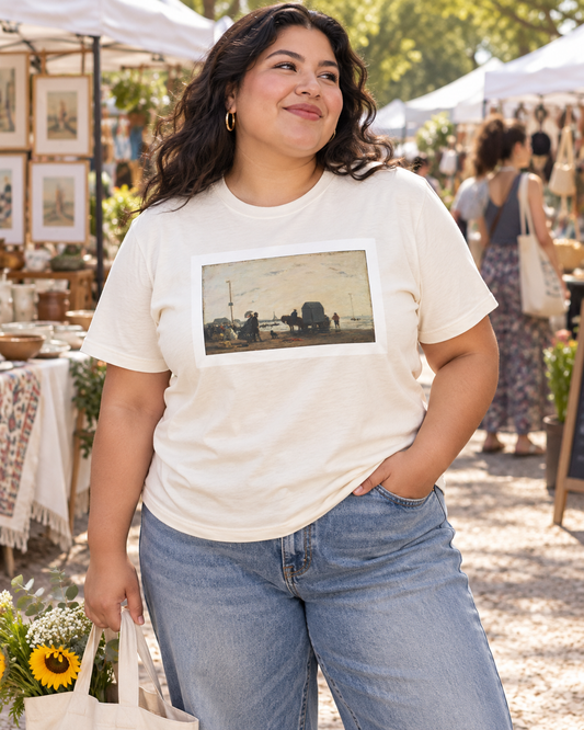 A woman wearing a cream-colored graphic t-shirt with a framed impressionist landscape print, paired with blue jeans, standing at an outdoor market.
