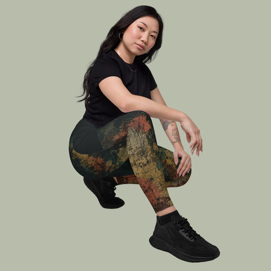 A woman crouching in black athletic leggings with a distressed multicolor print in earthy tones of green, brown, and orange, paired with a black t-shirt and black sneakers, against a plain white background.