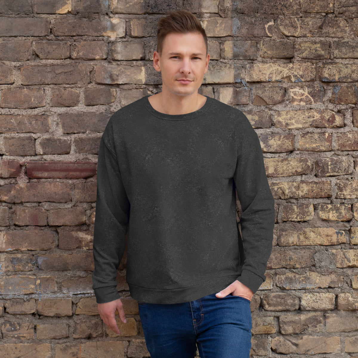 A man wearing a dark charcoal gray long-sleeve crewneck sweatshirt and blue jeans, standing against a rustic brick wall.