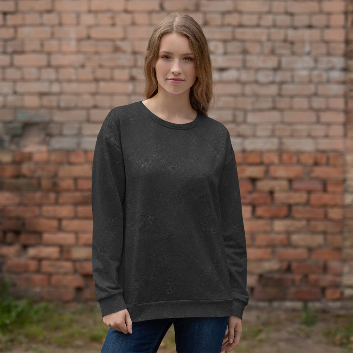 A woman wearing a dark charcoal gray long-sleeve crewneck sweatshirt and blue jeans, standing in front of a blurred red brick wall.