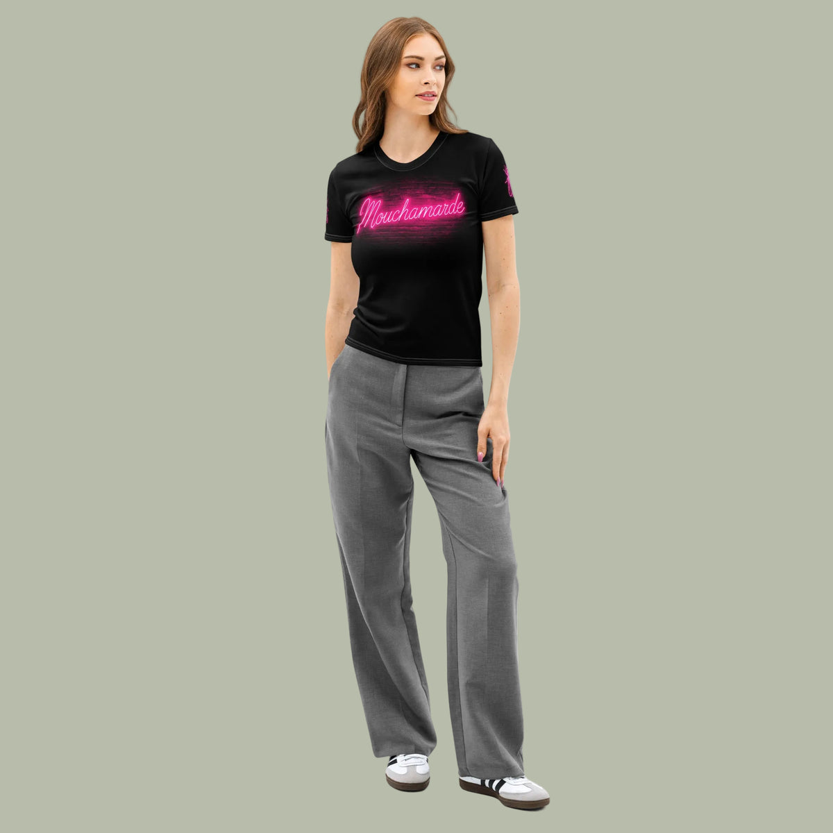 A woman modeling a black short-sleeve t-shirt with a pink 'Mouchamarde' logo on the chest, paired with grey wide-leg pants and white sneakers, against a plain sage background.