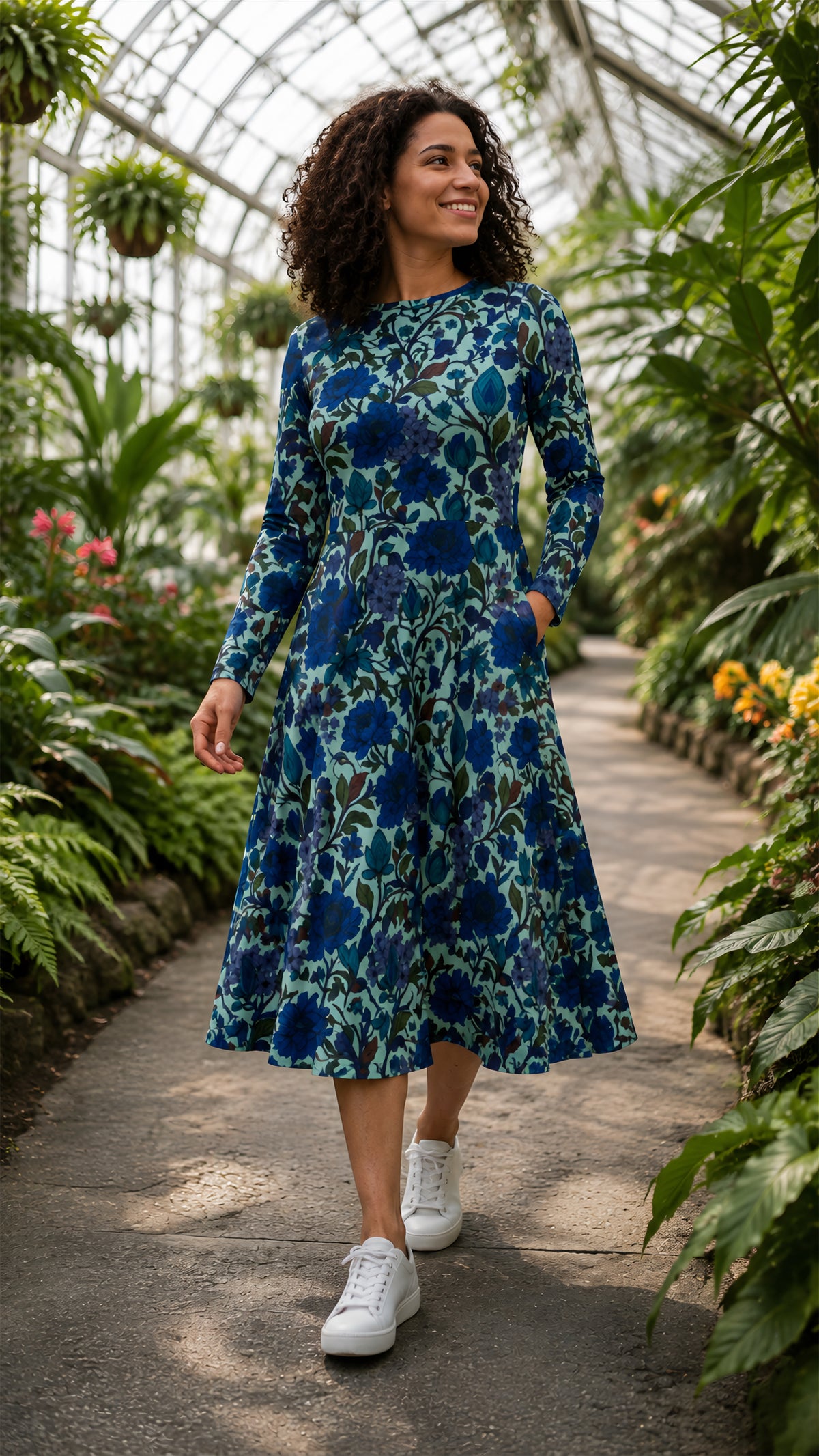 A woman wearing a long-sleeve midi dress with a blue and teal floral pattern walks along a stone path inside a lush green greenhouse.
