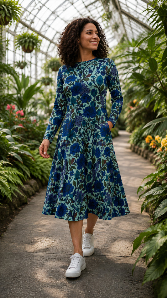 A woman wearing a long-sleeve midi dress with a blue and teal floral pattern walks along a stone path inside a lush green greenhouse.