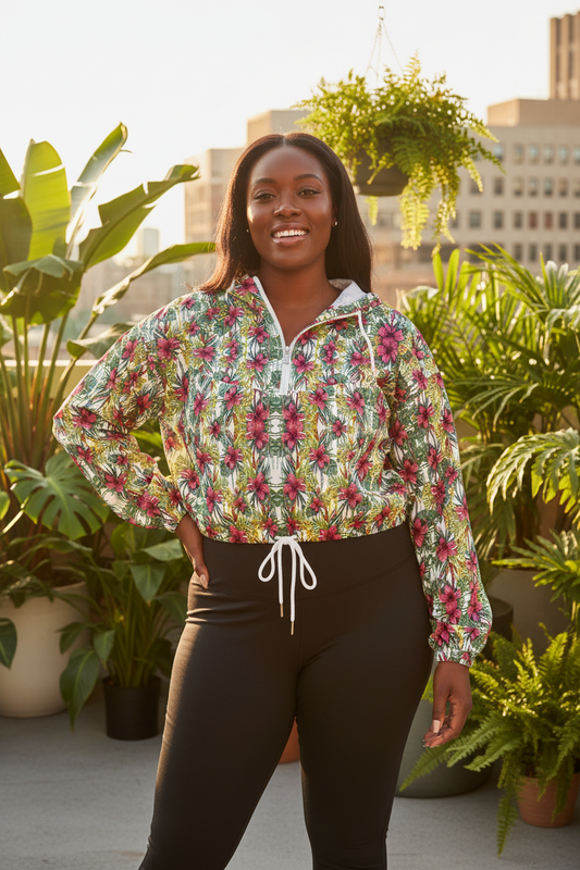 A woman stands on a rooftop wearing a Mint Blossom long-sleeve cropped windbreaker with a colourful tropical floral print and black leggings, surrounded by potted plants and buildings in the background.