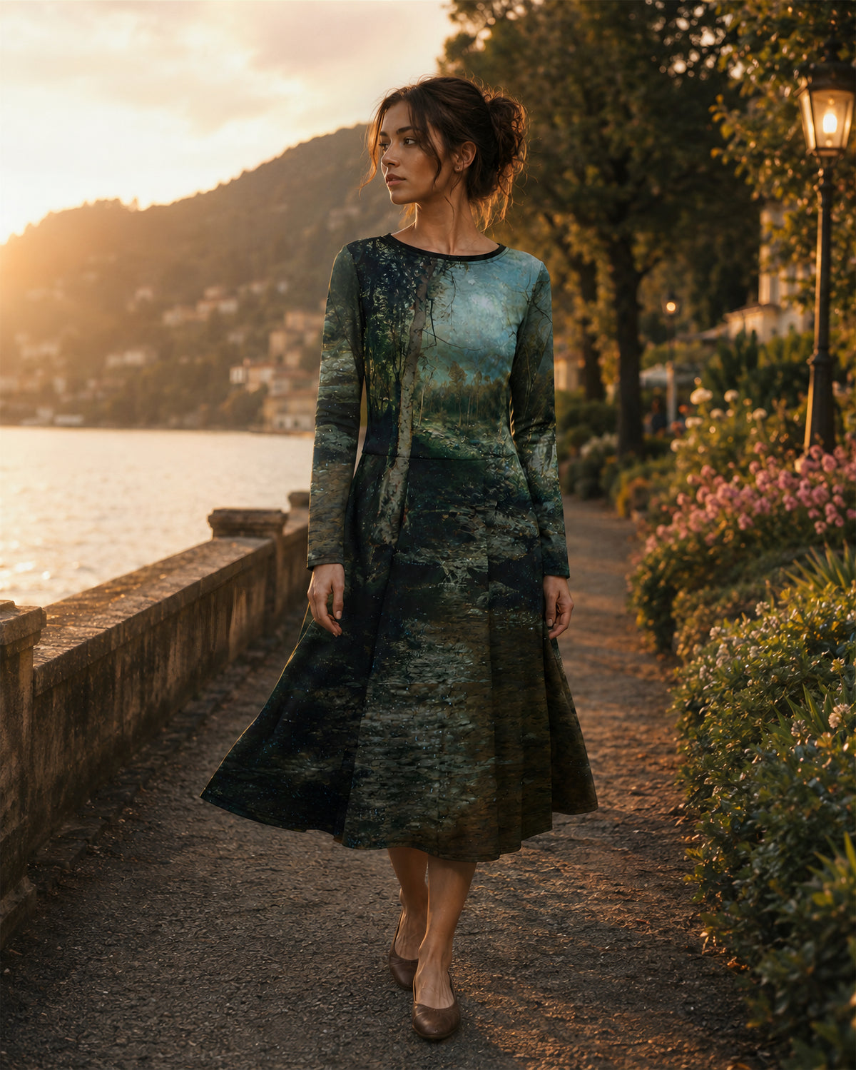 A woman wearing a long-sleeve midi dress with a scenic landscape print walks along a lakeside path at sunset. The dress features a dark green and blue watercolor-style print of trees and water, and she is wearing brown pointed-toe flats.