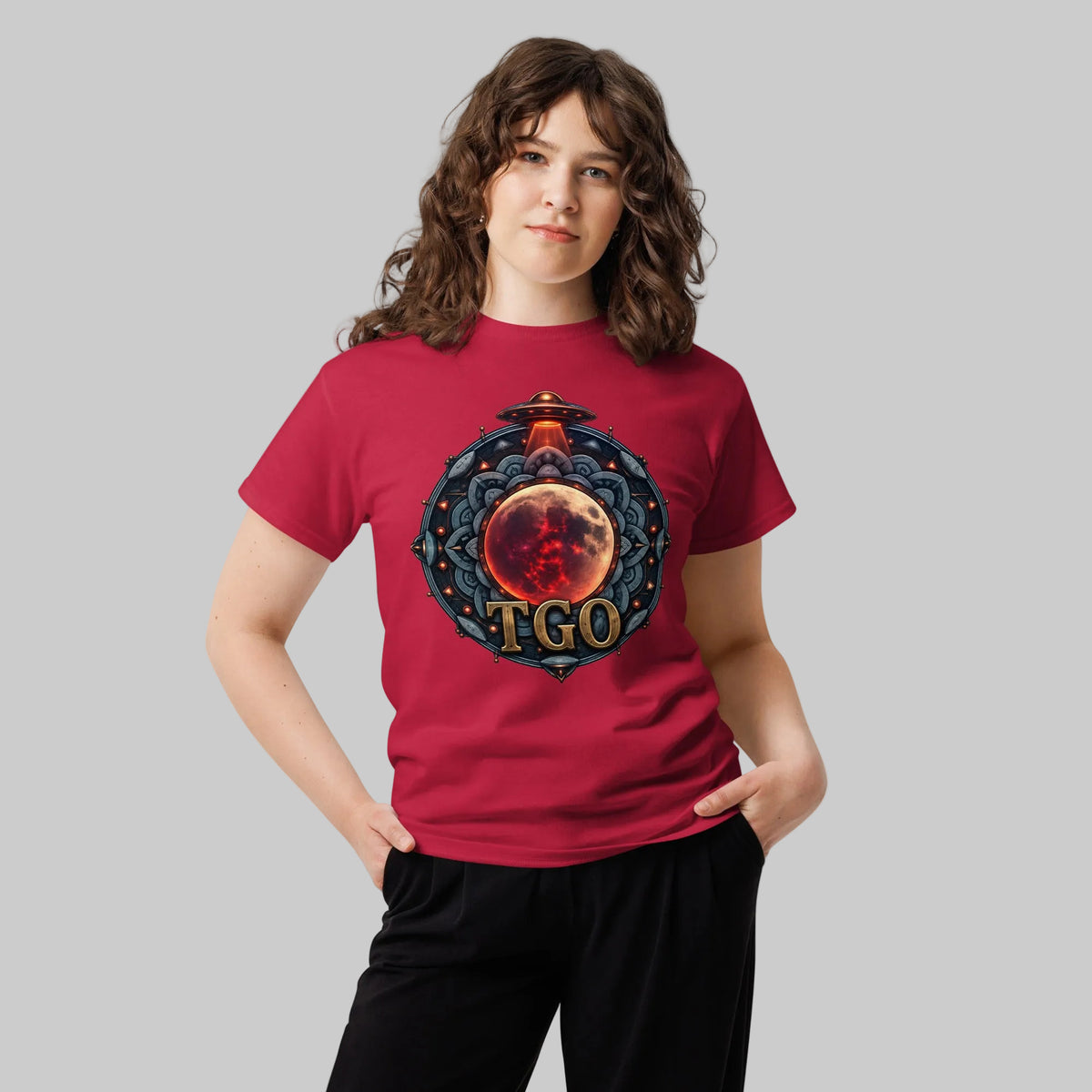 A woman with wavy brown hair wearing a red tshirt with a UFO Emblem and wearing black pants with a light grey background
